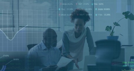Reviewing two colleagues in business wear leaning at office desk, pointing at laptop, paper, charts