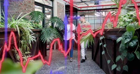 Walking colleagues in business attire discussing in office atrium holding tablet phone by planters