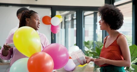 Decorating coworkers wearing pink top and red polka top in office lounge, holding balloons and gift