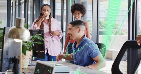 Smiling man wearing blue shirt using desktop monitor in open-plan office, showing green charts