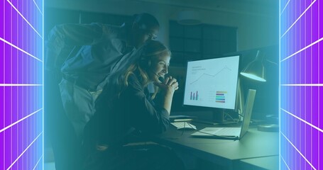 Analyzing two coworkers in shirts leaning over monitor at night office, with laptop and charts