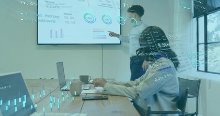 Pointing presenter in light shirt and dark pants presenting charts in meeting room, with display