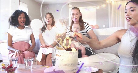 Celebrating five women sitting on light sofa, holding infant, watching gold '30' cake sparklers