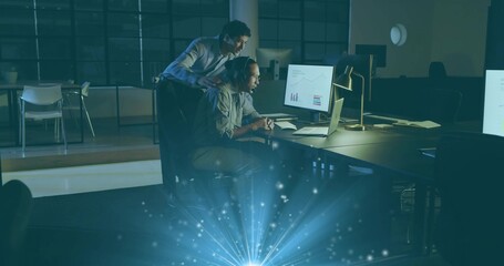 Reviewing coworkers in shirts leaning over laptop and monitor in open office with lamp, blue flare