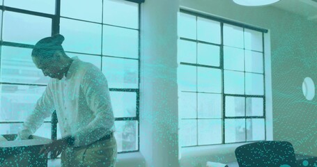 Leaning worker wearing light shirt, pants reviewing papers at desk by windows, with teal overlay