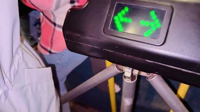 Bus payment gate indicator showing red X to green arrows, closeup of black plastic housing with led matrix, yellow pole and passenger in plaid shirt visible, urban transit