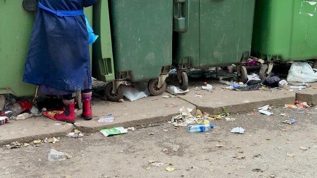 Elderly white woman rummaging dumpster in urban alley, worn blue coat and red boots, cane for support, blue plastic bag, scattered food wrappers and bottles, overflowing green
