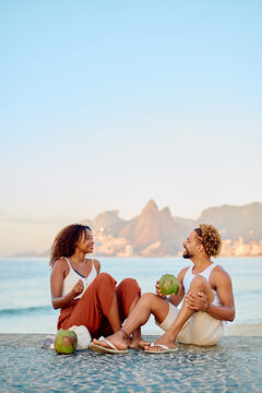 Young couple sitting, drinking fresh coconut water, relaxing on beach with city skyline and mountains in background