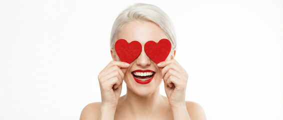 Happy blonde woman holding red glitter hearts over her eyes, smiling broadly on a white background, expressing love and joy.