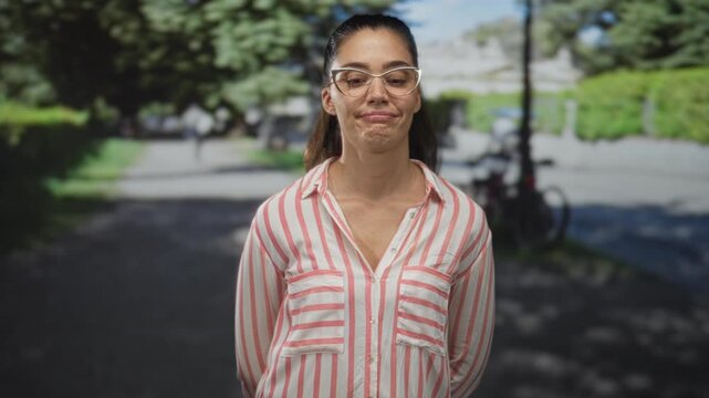 Woman with arms behind back wears glasses and striped shirt while standing in green park; skepticism doubt distrust cynicism.