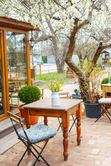 Charming spring garden scene with wooden table, chairs, and fresh flowers surrounded by blooming trees. Cozy outdoor cafe atmosphere with rustic furniture and natural seasonal landscape