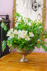 Elegant bouquet of white alstroemeria, peruvian lily flower in vintage bronze vase placed on wooden table with mirror and soft pastel interior background.
