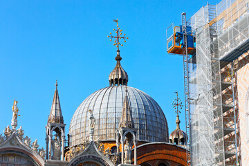 St Mark Basilica in Venice with its iconic domes and ornate details under clear blue sky. Scaffolding and construction lift show restoration work on the historic landmark