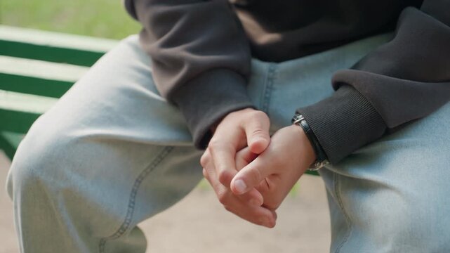 hands fidgeting on park bench closeup, person seated in denim jeans and dark sweatshirt, wristwatch visible, fingers rubbing in nervous rhythm, soft daylight and empty green bench slats, introspective