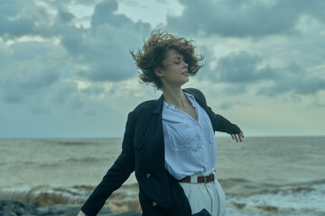 Wind, beach, ocean, movement, coat, shirt frame a windy scene as a woman on the shore stretches arms, faces the sea, and basks in the dramatic sky with rolling waves and misty light © SHOTPRIME STUDIO