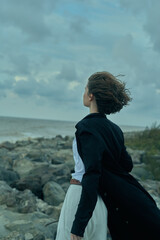 Fototapeta premium Woman standing on rocky coastline, wind blowing her hair, back turned, wearing a dress and dark jacket, contemplative mood as sea and sky meet at the horizon