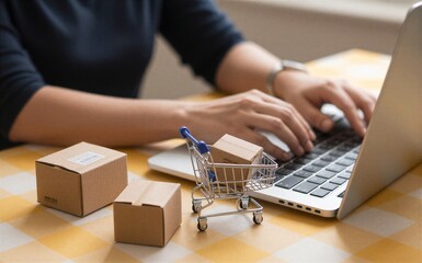 Person shops online, tiny shopping cart and boxes beside laptop on checkered table.