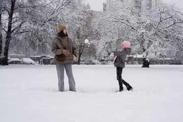 smiling girl in a pink hat throws a snowball at her laughing mother in a snowy winter park with trees and cars in the background joyful outdoor winter activity family fun recreation travel advertising