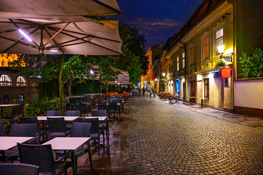 Illuminated sidewalk cafes and shops along the cobblestone Ljubljanica River embankment at night in the old town center of Ljubljana, Slovenia.