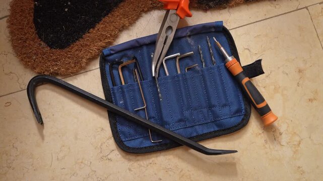 A set of lock picking tools, a crowbar, and pliers are spread on the floor near a blue tool roll and a doormat, indicating a break-in or burglary preparation scene