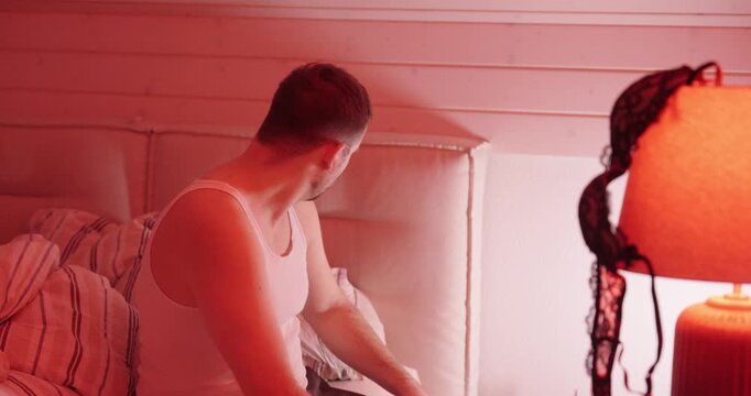 A sad, disappointed man sits on a bed in a red-lit room and slides his wedding ring back on, expressing guilt, remorse, and relationship conflict after an affair.