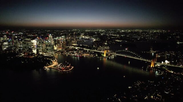 Drone high-angle aerial footage showing the illuminated Sydney Harbour Bridge and the glowing city skyline of North Sydney's business district at nigh