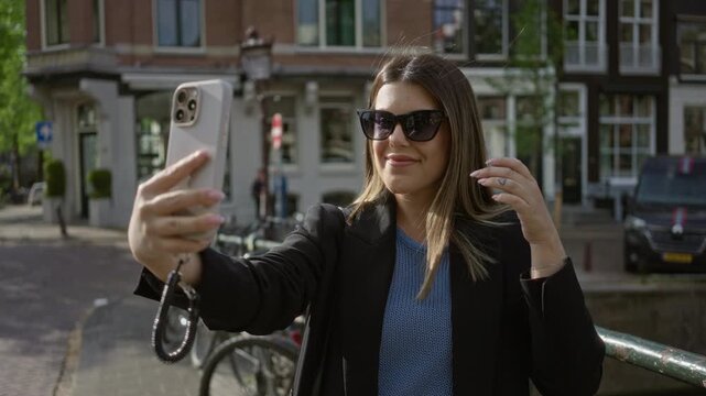 Woman taking selfie and smiling with smartphone in amsterdam, walking along scenic canals in stylish outfit during sunny day showcasing iconic dutch cityscape and vibrant atmosphere.