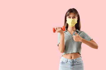 Young woman in medical mask with dumbbell showing thumb-up on pink background. Immunity concept