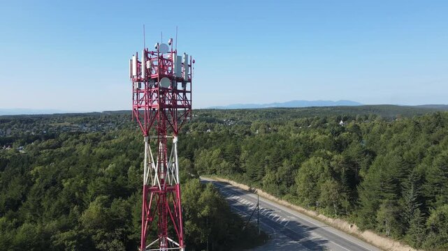 Aerial drone footage of a telecommunications tower standing above a forested area. Red and white metal structure with antennas providing mobile network and data transmission in a rural region. Highway