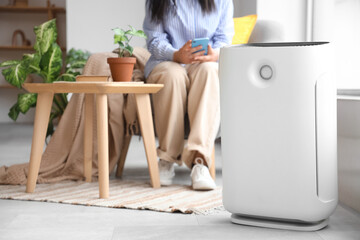 Woman in armchair adjusting air purifier using mobile phone at home
