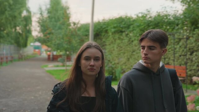 white teens walking along zoo path, backpacks and hoodies, stopping to read animal poster and glance at exhibit with hedged backdrop and red booth nearby cinematic evening light highlights curiosity,