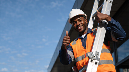 Positive construction worker in safety gear giving a thumbs up. Smiling African American builder holding a ladder outdoors with copy space for text