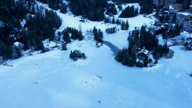 Aerial drone footage of a frozen lake in Champex, Switzerland, during winter. People ice skating surrounded by snowy alpine scenery. Shot in 4K using a DJI Mini 2.