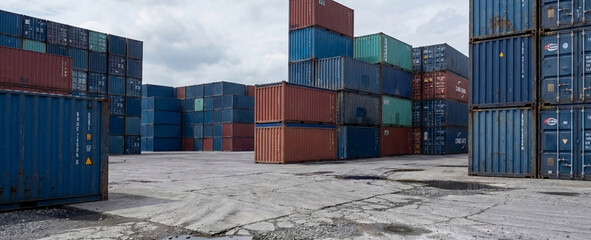 Colorful shipping containers are stacked in a large storage area, showcasing a combination of sizes and colors. The concrete ground is weathered, hinting at frequent use.