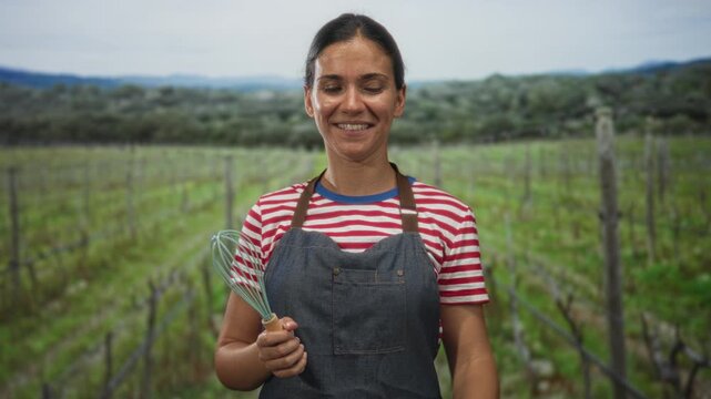 Woman wearing apron holding a whisk and making rock horns hand gesture in a vineyard field under open sky; rustic cooking joy.