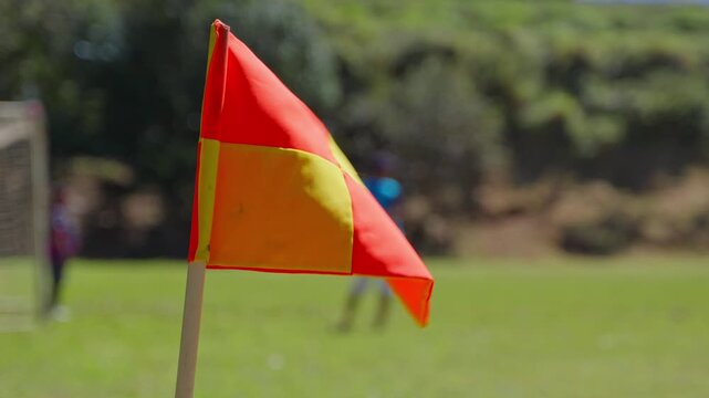 Detailed shot of a soccer corner flag waving in the wind on a green field, with a professional athlete performing drills in the background.