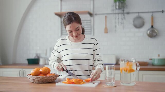 Cheerful young Asian woman smiling while slicing fresh oranges in a bright, modern kitchen, focusing on nutritious breakfast prep and a healthy longevity lifestyle at home.