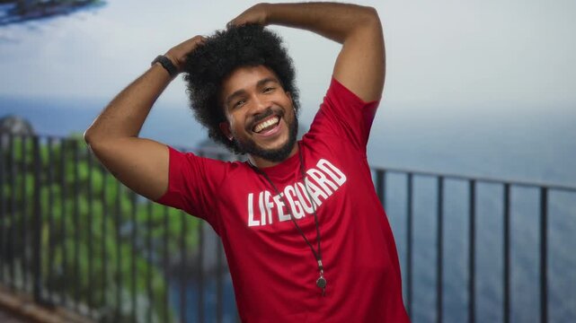 Lifeguard man smiling by the seaside promenade with heart gesture, wearing red uniform, against the scenic beach and sea outdoor backdrop.