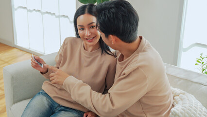 Asian man and woman feel excited while viewing an ultrasound image together on the sofa, sharing pregnancy news and an emotional relationship moment at home.