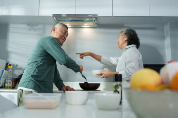 Asian elderly man and woman talk together while cooking in the kitchen, showing senior couple lifestyle with warm interaction and daily home activity.