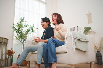 Obraz premium Young Asian lovers sit on the sofa cheering sports together, capturing a relaxed lifestyle moment with a man and woman enjoying their relationship at home.
