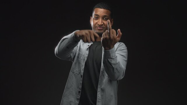 Young black man in gray shirt smiling while showing middle finger and pointing with hand in studio; defiance.