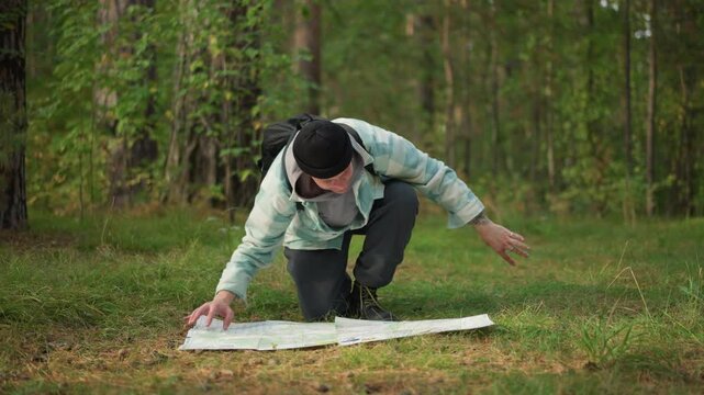 male hiker kneeling studying map, backpacked traveler examining routes on ground, forest clearing setting with trees and grass, focused route plotting and waypoint checking, sense of determination