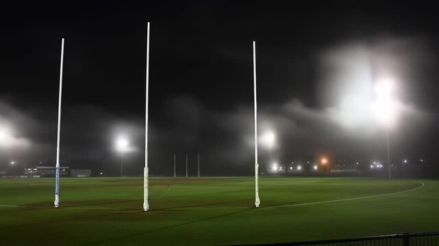 Foggy night scene of a sports field with goal posts illuminated by stadium lights.