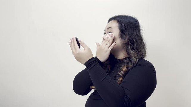 Young woman applying makeup with a compact mirror on a plain background, looking into the mirror and touching her face with a makeup applicator