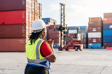 African female logistics manager in a hard hat using a walkie-talkie to coordinate container shipping operations at a busy industrial port terminal with reach stacker and cargo crates.