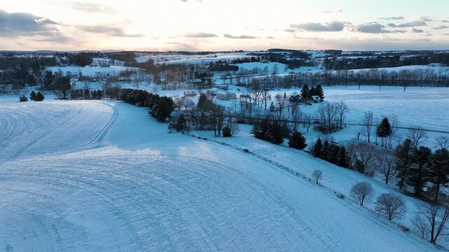 Aerial flyover snowy winter farmland fields at sunset in america. Wide shot. Car on intersection road. Dusk scene. Hills and countryside of rural area.