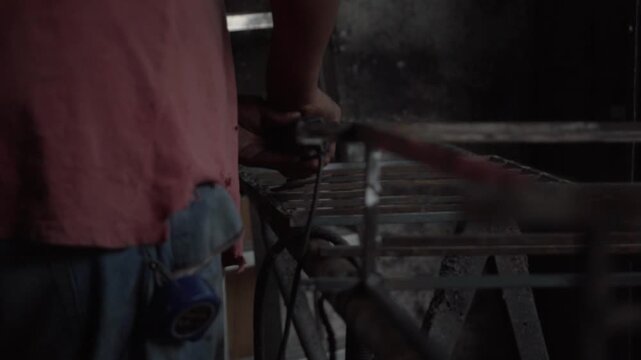 Worker grinding metal bars with angle grinder as sparks fly inside an industrial workshop.