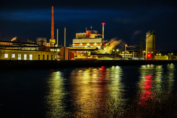 The Gersthofen industrial park at night. Industrial site with several buildings and tall chimneys, from which steam rises into the night sky. Reflection of artificial lighting in the Lech Canal.