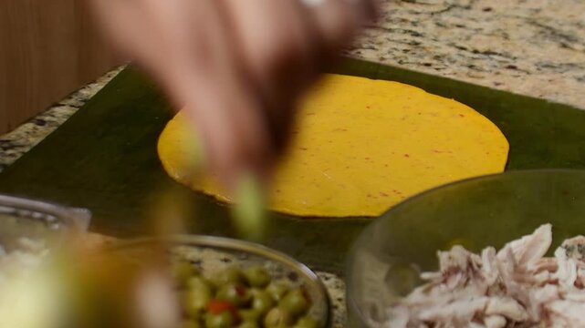 Close-up of hands assembling hallacas on a kitchen table, traditional Venezuelan dish, folding masa and fillings into palm leaves, festive homemade cooking.